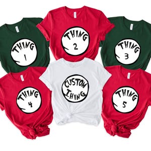 May include: A collection of short-sleeved t-shirts in red, white, and green. Each shirt features a circular graphic with the word "THING" and a number or custom text. The shirts are arranged in a group, suitable for themed events.
