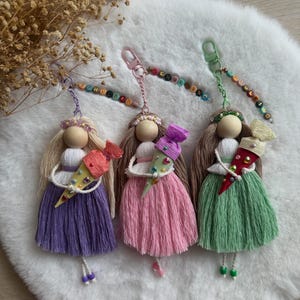 May include: Three handmade doll keychains with wooden bead heads and yarn hair. Each doll wears a dress in purple, pink, or green and holds a small cone-shaped gift bag. The keychains feature colorful letter beads spelling SCHULTÜTE and metal clips.