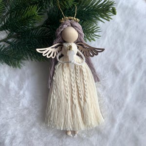 Könnte beinhalten: Handgefertigter Engelsornament mit Holzkopf, lavendelfarbenem Haar und goldfarbenen Flügeln. Der Engel trägt ein cremefarbenes Kleid und hält einen Kristall. Ein goldfarbener Heiligenschein und eine Aufhängeschnur vervollständigen das Design. Der Ornament ist ca. 15 cm hoch.