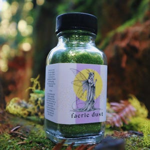 May include: A clear glass bottle filled with green glitter, labeled "faerie dust." The label features a fairy illustration with a yellow circle background. The bottle has a black cap and sits on a mossy surface.