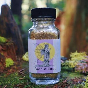 May include: A clear glass bottle filled with gold glitter, labeled "faerie dust." The label features a fairy illustration with a yellow moon background. The bottle has a black cap and is set against a blurred natural background.