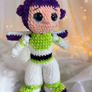 May include: A handmade crochet doll of a cartoon character, featuring a purple head and accents, a light pink face, and large blue eyes. The doll wears a green and white outfit with white arms and legs. The doll is approximately 10 inches tall.