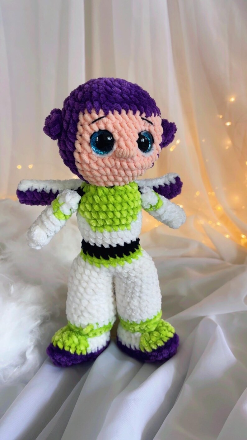 May include: A handmade crochet doll of a cartoon character, featuring a purple head and accents, a light pink face, and large blue eyes. The doll wears a green and white outfit with white arms and legs. The doll is approximately 10 inches tall.