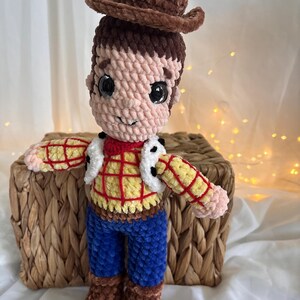 May include: A handmade crochet doll of a cowboy character, featuring a brown hat, brown hair, and a plaid shirt with a white vest. The doll has blue pants and brown boots. The doll is sitting on a woven basket.