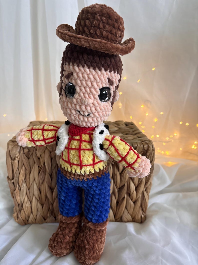 May include: A handmade crochet doll of a cowboy character, featuring a brown hat, brown hair, and a plaid shirt with a white vest. The doll has blue pants and brown boots. The doll is sitting on a woven basket.