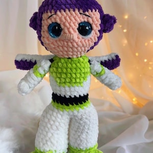 May include: A handmade crochet doll of a character with a purple head, pink face, and large blue eyes. The doll wears a white and green outfit with purple accents. The doll is approximately 10 inches tall.