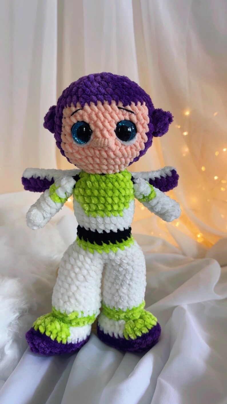 May include: A handmade crochet doll of a character with a purple head, pink face, and large blue eyes. The doll wears a white and green outfit with purple accents. The doll is approximately 10 inches tall.