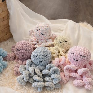 May include: A collection of handmade crochet octopus plushies in various pastel colors, including pink, blue, purple, and white. Each octopus has a smiling face and curled tentacles, resting on a soft, fluffy surface.