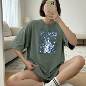 May include: A sage green short-sleeve t-shirt featuring a blue graphic print on the front. The design depicts the Statue of Liberty, the text USA, the number 250, and several stars arranged within a rectangular border on the chest area.