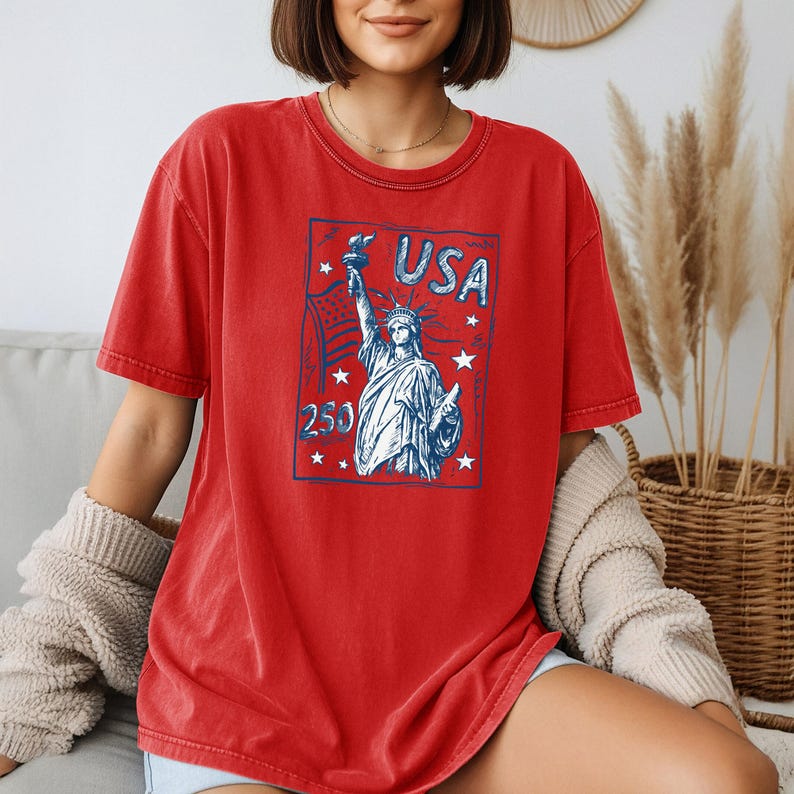 May include: A red short-sleeved t-shirt featuring a blue graphic print of the Statue of Liberty, the text USA, the number 250, and several stars. The design is enclosed within a rectangular border, creating a vintage-inspired patriotic aesthetic.