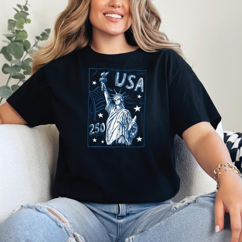 May include: A black short-sleeved t-shirt featuring a blue graphic print of the Statue of Liberty, the text USA, the number 250, and several stars. The design is framed within a rectangular border, showcasing a patriotic theme on the front of the shirt.