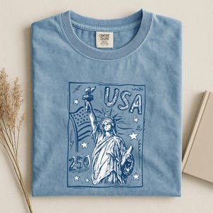 May include: A blue crew neck t-shirt featuring a dark blue graphic print of the Statue of Liberty, an American flag, stars, and the text USA 250. The shirt has a Comfort Colors brand label on the inside of the collar and a short-sleeved design.