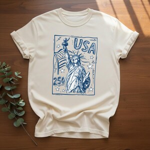 May include: A cream-colored short-sleeve t-shirt featuring a blue graphic print of the Statue of Liberty, an American flag, and stars. The design includes the text USA and the number 250, styled like a postage stamp with a sketch-like appearance.