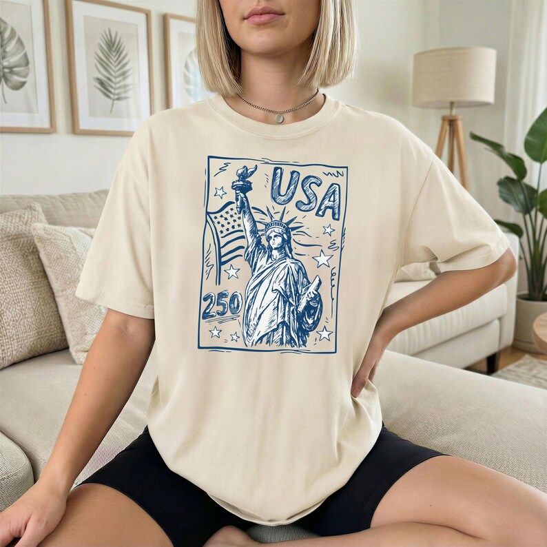 May include: A beige short-sleeve t-shirt featuring a blue graphic print of the Statue of Liberty, an American flag, stars, and the text USA 250. The design is enclosed in a rectangular border, centered on the front of the crew neck garment.