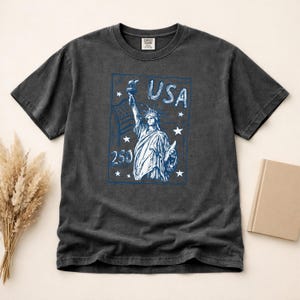 May include: A charcoal gray short-sleeve t-shirt featuring a blue graphic print of the Statue of Liberty, the text USA, the number 250, and several stars. The shirt has a crew neckline and a label at the inner collar area with visible text.