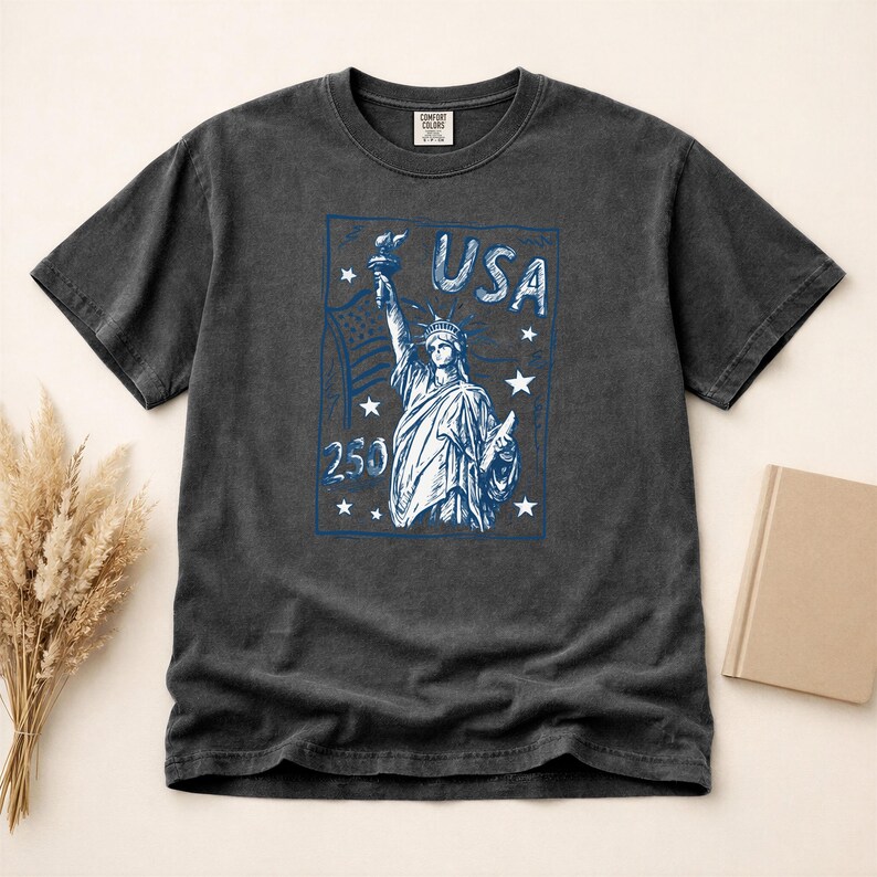 May include: A charcoal gray short-sleeve t-shirt featuring a blue graphic print of the Statue of Liberty, the text USA, the number 250, and several stars. The shirt has a crew neckline and a label at the inner collar area with visible text.