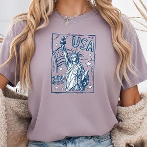 May include: A light purple t-shirt featuring a dark blue graphic print of the Statue of Liberty, the American flag, stars, and the text USA and 250. The design is enclosed in a rectangular border with a sketch-like artistic style.