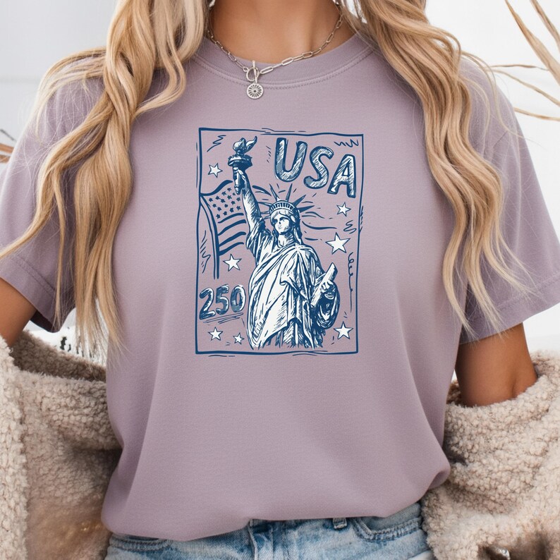 May include: A light purple t-shirt featuring a dark blue graphic print of the Statue of Liberty, the American flag, stars, and the text USA and 250. The design is enclosed in a rectangular border with a sketch-like artistic style.