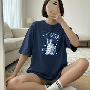 May include: A navy blue short-sleeved t-shirt featuring a white graphic print of the Statue of Liberty, the text USA, and the number 250, surrounded by stars and a rectangular border. The shirt has a crew neckline and a relaxed, loose-fitting cut.