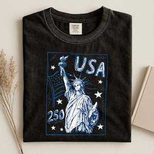 May include: A black crew neck t-shirt featuring a blue and white graphic print of the Statue of Liberty, the American flag, stars, and the text "USA" and "250". The shirt has a Comfort Colors brand label on the inside of the collar.