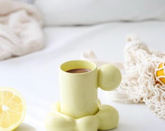 Puede incluir: Una taza de cerámica amarillo pálido con un asa y una base redondeadas únicas, llena de una bebida oscura. La taza está sobre una superficie blanca, junto a un limón a la mitad y una revista. El diseño de la taza es moderno y artístico.