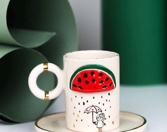 Puede incluir: Taza de cerámica blanca con diseño de sandía y platillo a juego. La taza presenta una rodaja de sandía roja, gotas de lluvia y una niña con un paraguas. El asa tiene detalles dorados. El platillo tiene un borde dorado.