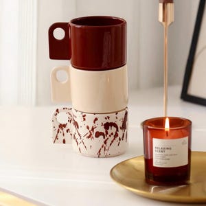 May include: Three stacked ceramic mugs in burgundy, cream, and cream with a burgundy splatter pattern. A lit candle in a glass jar sits on a gold-coloured tray. The candle label reads "Relaxing Scent".