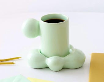 Puede incluir: Una taza de café de cerámica verde claro llena de líquido oscuro, con un diseño único con formas redondeadas. La taza está sobre una superficie blanca con artículos de papelería, incluyendo sobres y un cuaderno.