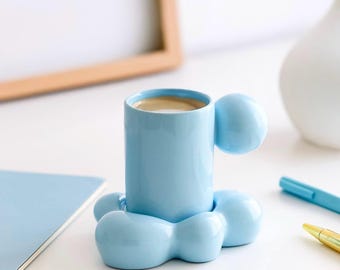 Puede incluir: Una taza de café de cerámica azul claro con un diseño único. La taza tiene un cuerpo cilíndrico, un asa redondeada y una base con tres soportes redondeados. La taza está llena de café. La taza está sobre una superficie blanca.