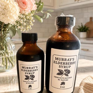 Peut inclure: Deux bouteilles en verre brun de sirop de sureau Murray's avec des bouchons noirs et des étiquettes blanches. Les étiquettes indiquent "MURRAY'S ELDERBERRY SYRUP" et "IMMUNITY REFINED BY NATURE". Une bouteille est étiquetée 4 oz, l'autre 8 oz.