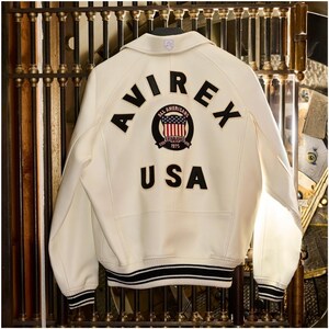 Op de afbeelding: Cr&egrave;mekleurige leren jas met het woord "AVIREX" en "USA" in zwarte letters op de rug. Een cirkelvormige patch met de woorden "All Americans Established 1975" en de Amerikaanse vlag is gecentreerd tussen de woorden. De jas heeft zwart-witte gestreepte afwerking.