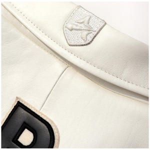 Op de afbeelding: Close-up van een witte leren jas met een gestikte naad. De jas heeft twee geborduurde patches: een wit embleem en de letter "D" in zwart en wit. Het leer heeft een gladde textuur.