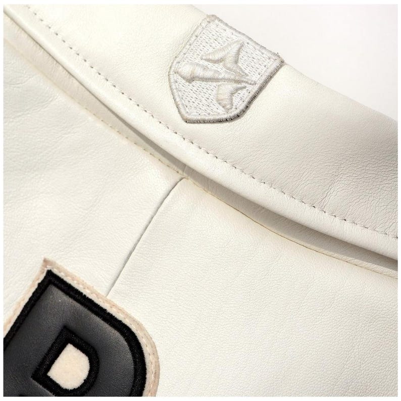 Op de afbeelding: Close-up van een witte leren jas met een gestikte naad. De jas heeft twee geborduurde patches: een wit embleem en de letter "D" in zwart en wit. Het leer heeft een gladde textuur.