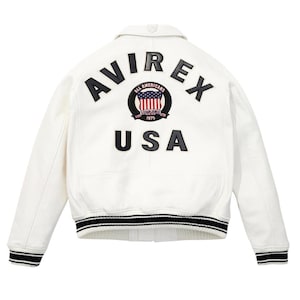 Op de afbeelding: Witte leren jas met zwart-wit gestreepte manchetten en kraag. De achterkant heeft het woord "AVIREX" boven een cirkelvormig embleem met de woorden "ALL AMERICANS ESTABLISHED 1975" en een USA-vlagontwerp. Daaronder staat het woord "USA".