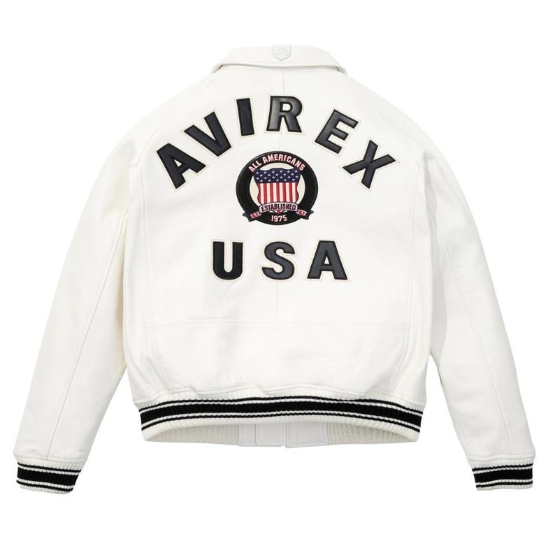 Op de afbeelding: Witte leren jas met zwart-wit gestreepte manchetten en kraag. De achterkant heeft het woord "AVIREX" boven een cirkelvormig embleem met de woorden "ALL AMERICANS ESTABLISHED 1975" en een USA-vlagontwerp. Daaronder staat het woord "USA".