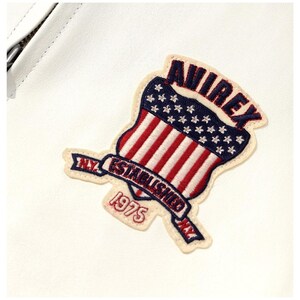 Op de afbeelding: Geborduurde patch met het woord "AVIREX" in rood en blauw, met een schildontwerp met rode en witte strepen en blauwe sterren. De patch bevat de tekst "N.Y. ESTABLISHED 1975" op een banner.