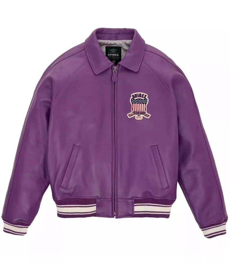 以下が含まれることがあります： 紫色の革製ボンバージャケット。襟とジッパー開閉部が付いています。胸にはアメリカ国旗のデザインとブランド名が施されたパッチが付いています。袖口と裾には白とクリーム色のストライプが入っています。