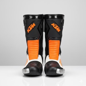Könnte beinhalten: Paar schwarze und orangefarbene Motorradstiefel mit dem KTM-Logo. Die Stiefel haben einen weißen Knöchelbereich und silberne Akzente an den Zehen. Entwickelt für Schutz und Leistung.