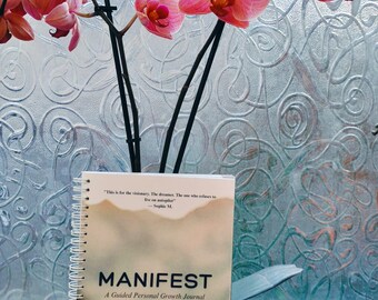 May include: A spiral-bound journal titled "MANIFEST: A Guided Personal Growth Journal" with a landscape design. The journal is placed in front of a window with a floral design, and a pink orchid plant with dark stems.