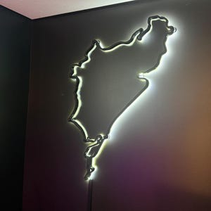 Pode incluir: Um contorno iluminado, montado na parede, de uma massa de terra, provavelmente um estado ou região. A luz é branca brilhante, criando um efeito brilhante contra a parede escura. A estética geral é moderna e minimalista.