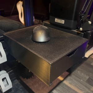 以下が含まれることがあります： 黒いコンピューターマウスが、引き出し付きの黒いテクスチャ加工されたトレイの上に置かれています。 トレイはゲーム設定の一部で、背景にはレーシングシミュレーターのペダルセットが見えます。