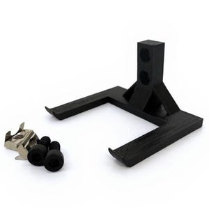 May include: A black, 3D-printed stand with a rectangular base and a vertical support featuring two holes. A small metal clip and several black rubber feet are positioned nearby. The stand is designed to hold an item.