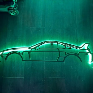 Puede incluir: Contorno de coche verde neón, iluminado sobre un fondo oscuro. El diseño del coche presenta un alerón y un perfil elegante. Un pequeño coche de juguete es visible en la esquina superior izquierda. El tema general es automotriz.