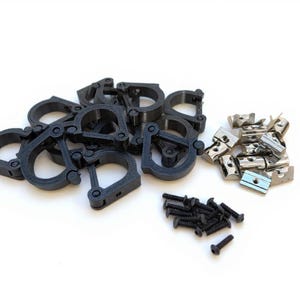 Könnte beinhalten: Eine Auswahl an Hardware-Komponenten, darunter schwarze Kunststoffklemmen, silberne Metallhalterungen und schwarze Schrauben, ist auf einer weißen Oberfläche angeordnet. Die Klemmen haben ein kreisförmiges Design mit einem Scharnier.