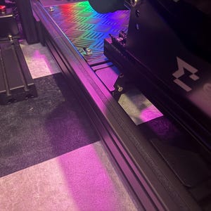 Puede incluir: Primer plano de un juego de pedales y un marco de simulador de carreras negros. El marco es de metal negro y tiene un reposapiés con diseño de diamante. Los pedales son negros con detalles plateados. La imagen está iluminada con luces moradas y arcoíris.