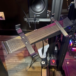 Op de afbeelding: Een zwarte en zilveren tablethouder met een geribbeld, houtachtig oppervlak. De houder is gemonteerd op een zwart frame, met een luidspreker en andere elektronische componenten op de achtergrond. De houder heeft een stevig ontwerp.