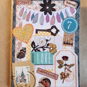Puede incluir: Una cubierta de cuaderno rectangular decorada con un collage de recortes de papel que incluye una guirnalda de borlas, una flor de metal oscuro, un corazón amarillo, una llave, un círculo azul con el número 7, una rosa, un pájaro y un sello alemán.