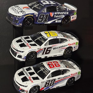 May include: Three miniature NASCAR race cars. The top car is navy blue and white with the number 91. The middle car is white with the number 16. The bottom car is white with the number 88. All cars have sponsor logos and Goodyear tires.