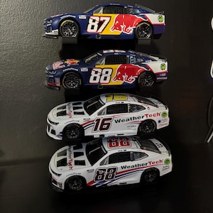 May include: Four toy race cars are displayed. Two are blue with Red Bull logos and the numbers 87 and 88. The other two are white with the WeatherTech logo and the numbers 16 and 88. All have Goodyear tires.