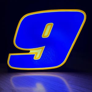 May include: A glowing blue number nine with a yellow outline and interior details. The number is illuminated against a dark background, casting a reflection on the wooden surface below. The design is modern and eye-catching.